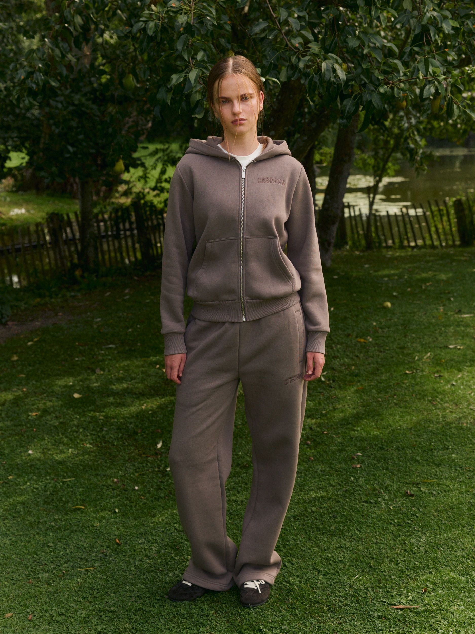 Woman wearing walnut straight-leg sweatpants and matching zip-up hoodie standing on a green lawn.